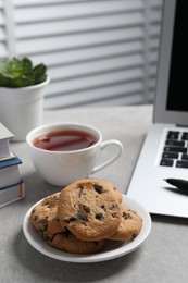 Photo of Chocolate chip cookies, cup of tea and laptop on light grey table in office