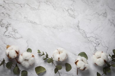 Fluffy cotton flowers and green leaves on white marble background, flat lay. Space for text