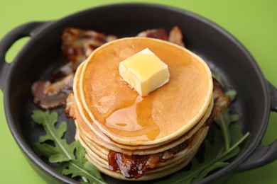 Photo of Tasty pancakes with butter, fried bacon and fresh arugula on light green background, closeup