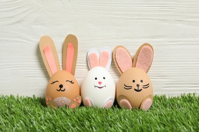 Easter eggs as cute bunnies on green grass against white wooden background