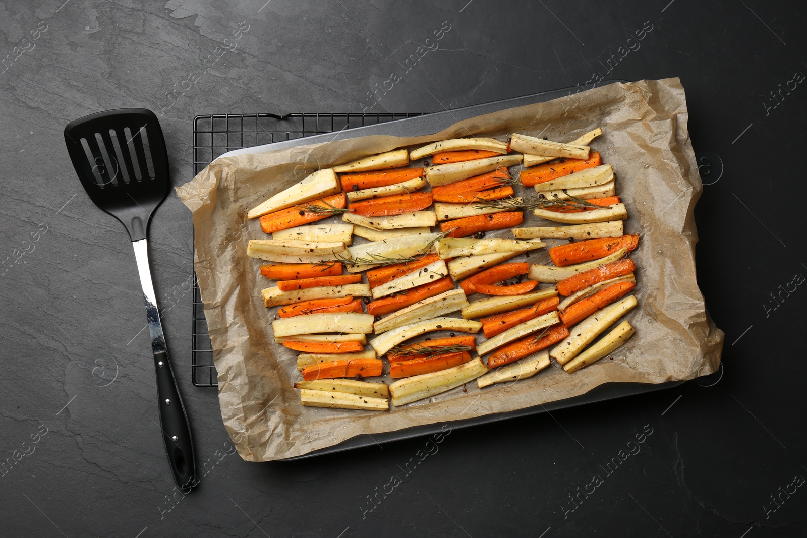 Photo of Tray with baked parsnips, carrots and spatula on black table, flat lay