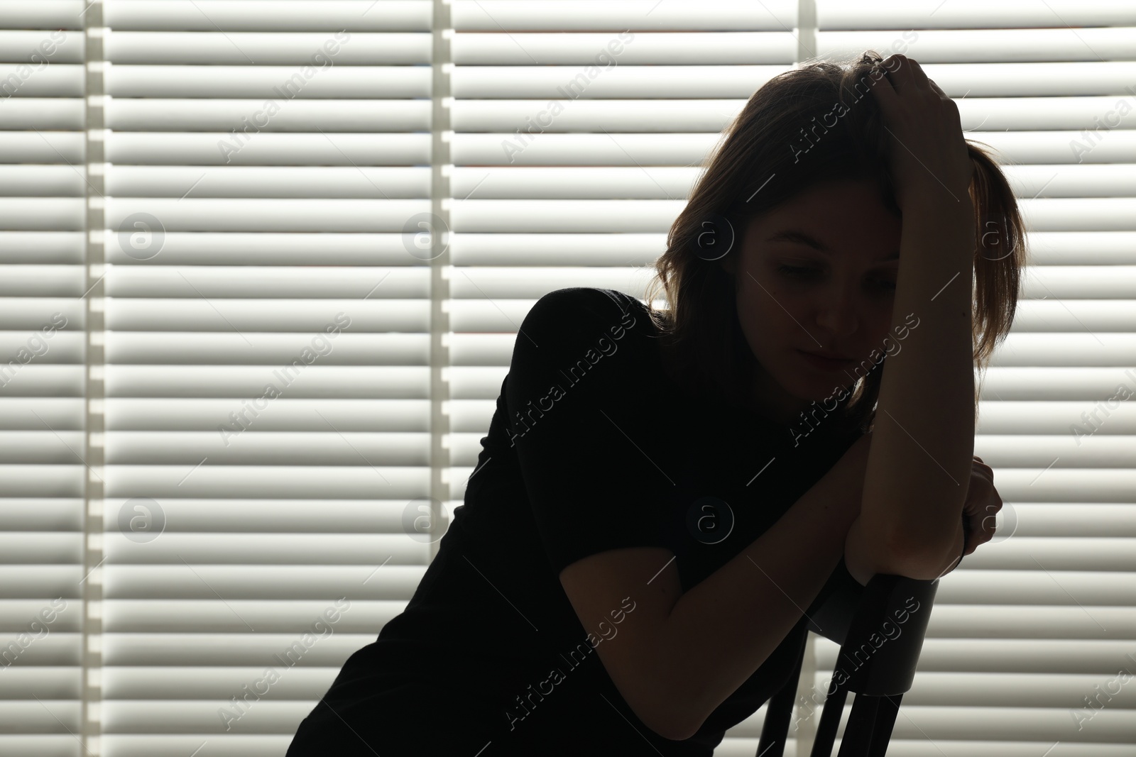 Photo of Silhouette of sad young woman near closed blinds indoors