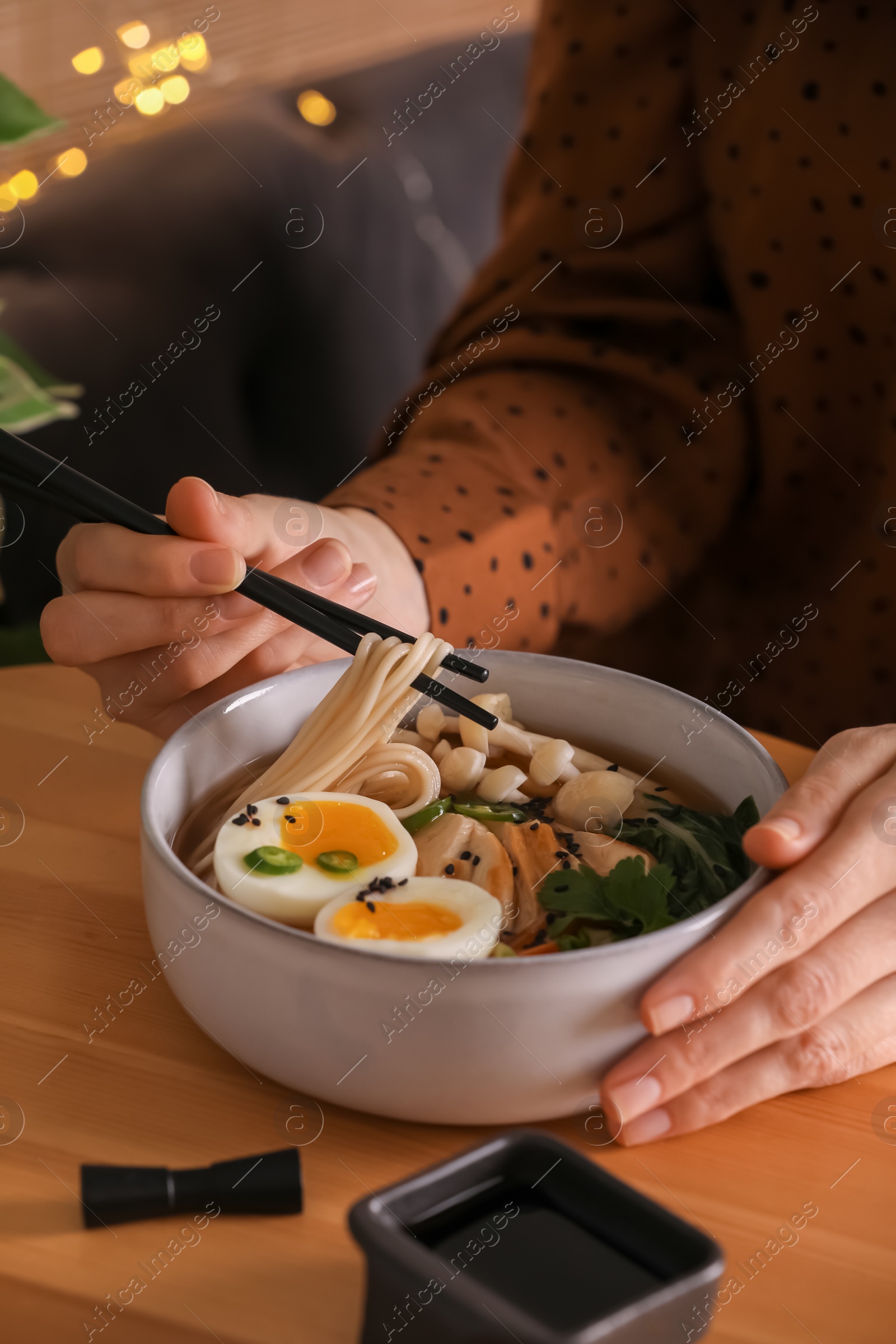 Photo of Woman eating delicious ramen with chopsticks at wooden table indoors, closeup. Noodle soup