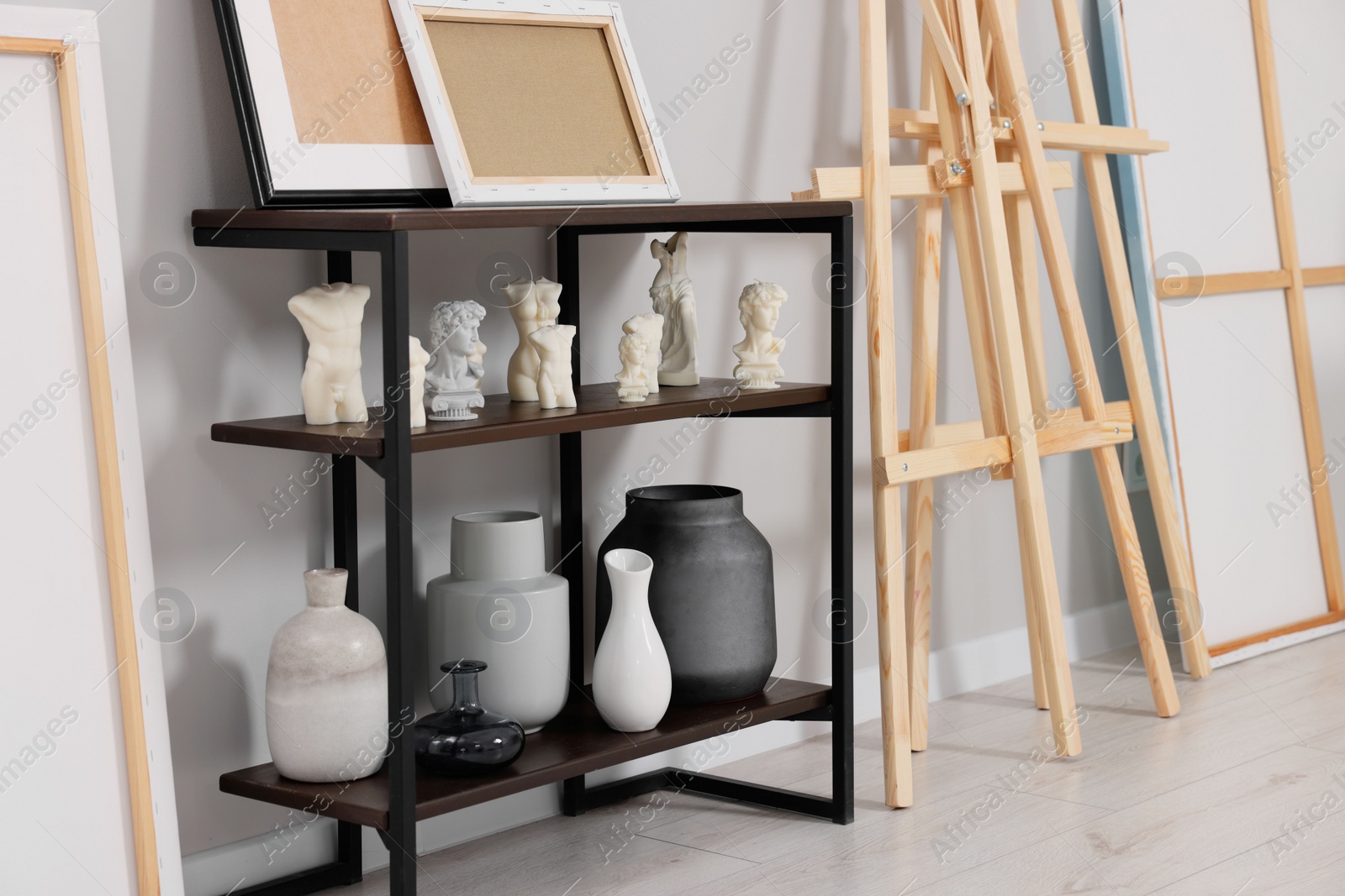 Photo of Wooden easel near shelving unit with canvases, vases and small sculptures in artist's studio