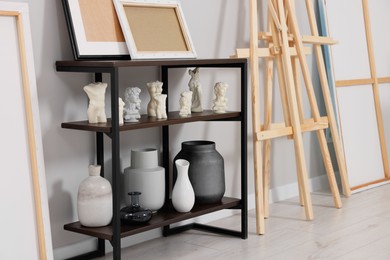 Photo of Wooden easel near shelving unit with canvases, vases and small sculptures in artist's studio