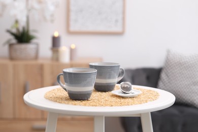 Photo of Cups of drink and snap infuser with dry leaves on white coffee table at home
