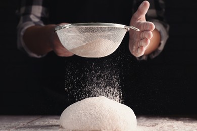 Man sprinkling flour over dough at wooden table on dark background, closeup
