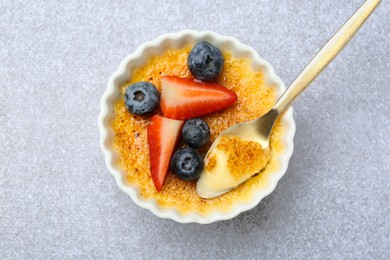 Photo of Delicious creme brulee with berries in bowl and spoon on grey textured table, top view