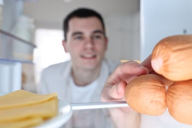 Photo of Happy man taking sausages out of refrigerator in kitchen, view from inside
