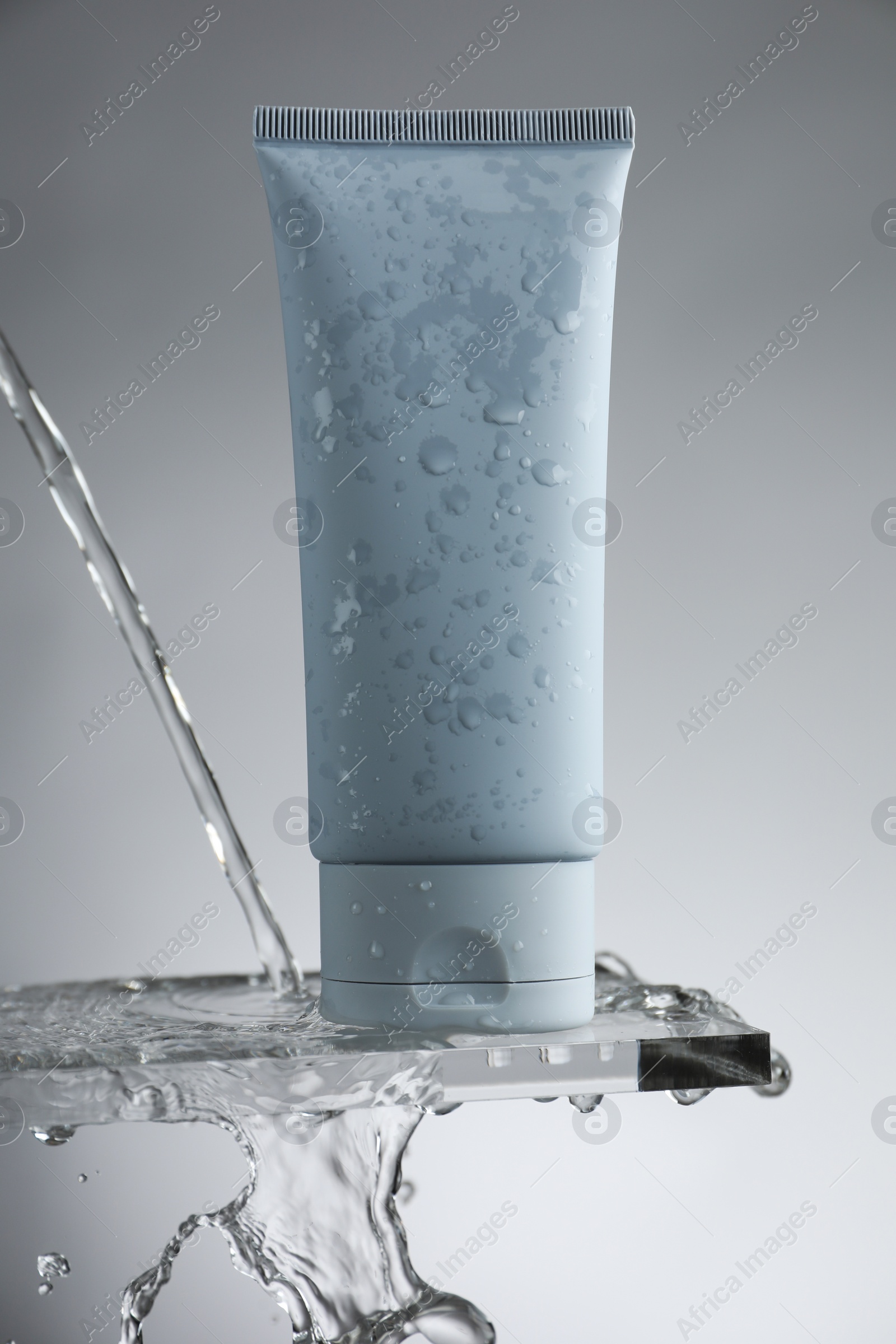 Photo of Moisturizing cream in tube on glass with water drops against grey background