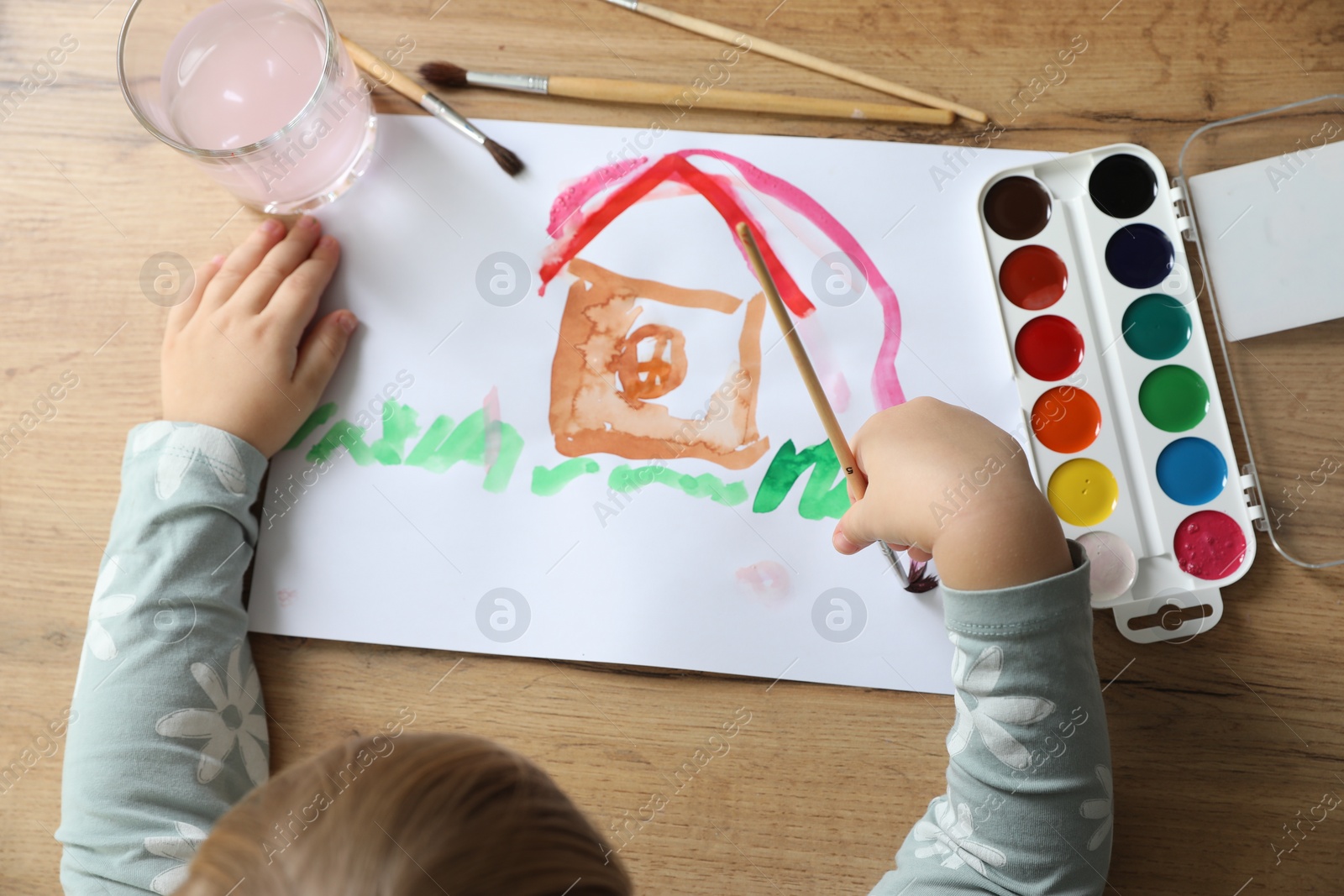 Photo of Cute little girl drawing house with brush at wooden table, top view. Child`s art