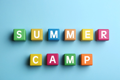 Colorful wooden cubes with phrase SUMMER CAMP on light blue background, flat lay