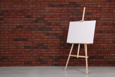 Photo of Wooden easel with blank canvas near brick wall indoors. Space for text