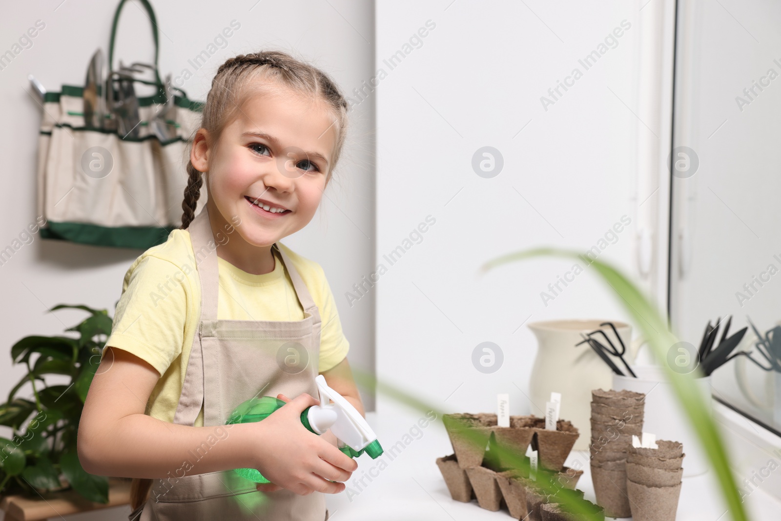 Photo of Little girl spraying water onto vegetable seeds in peat pots on window sill indoors