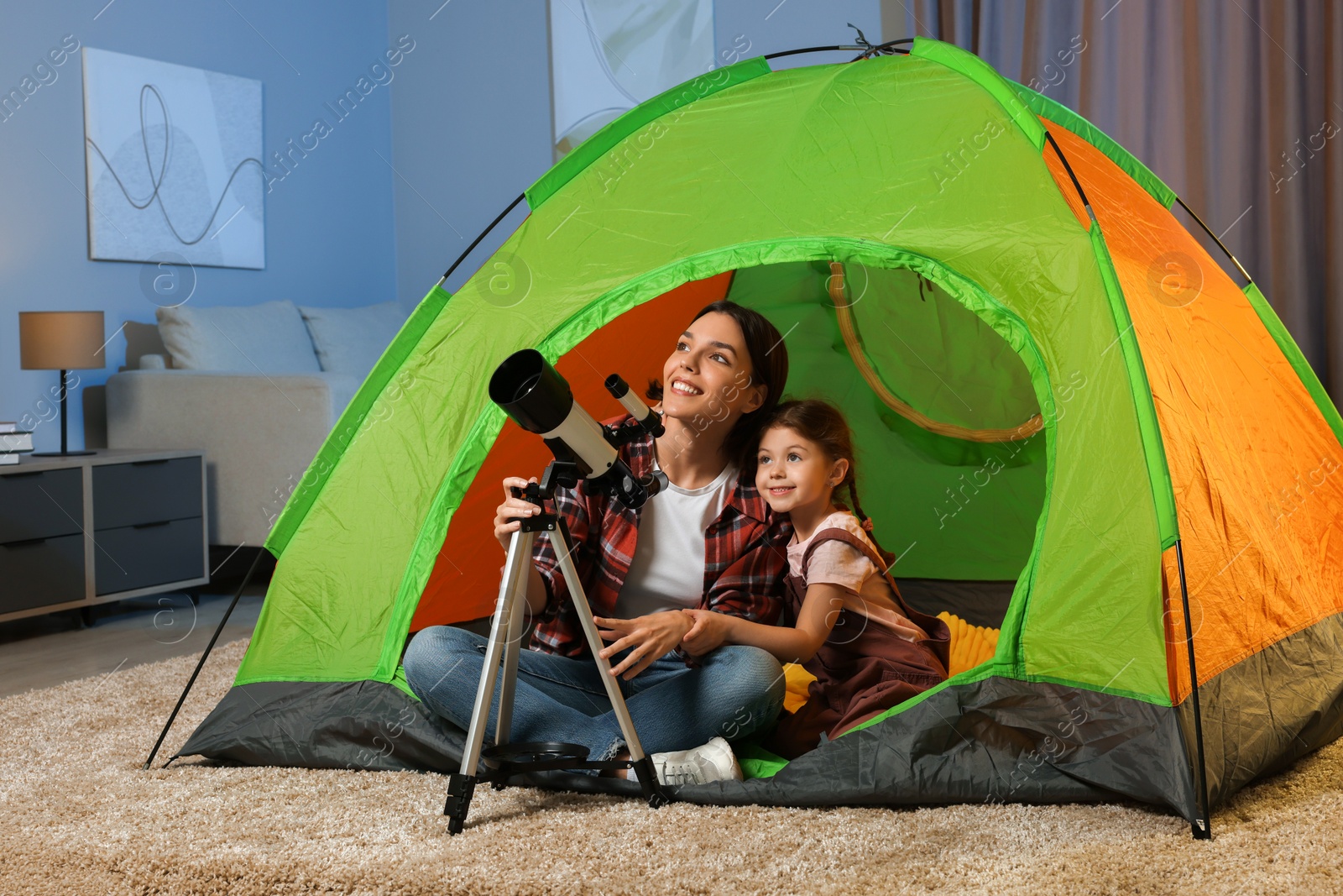 Photo of Happy mother and her daughter using telescope to look at stars while sitting in camping tent indoors