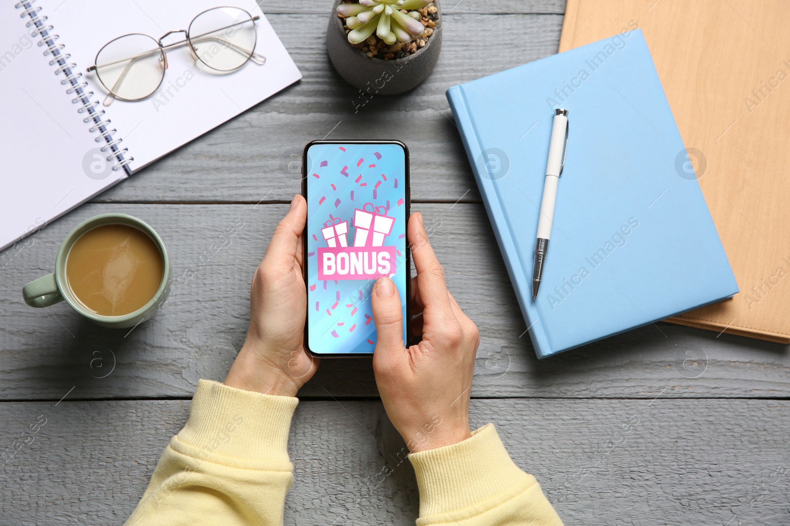 Image of Bonus gaining. Woman using smartphone at grey wooden table, top view. Illustration of gift boxes, word and falling confetti on device screen