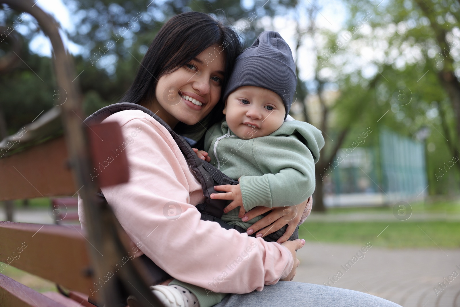 Photo of Mother holding her child in sling (baby carrier) on bench in park