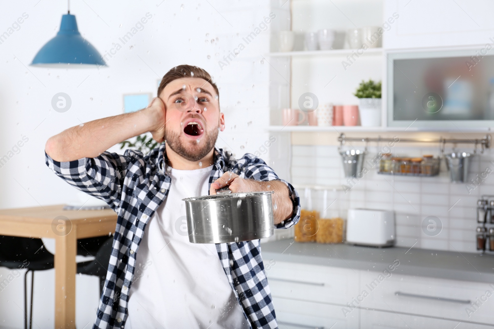 Photo of Emotional young man holding saucepan under water leakage from ceiling in kitchen, space for text. Plumber service