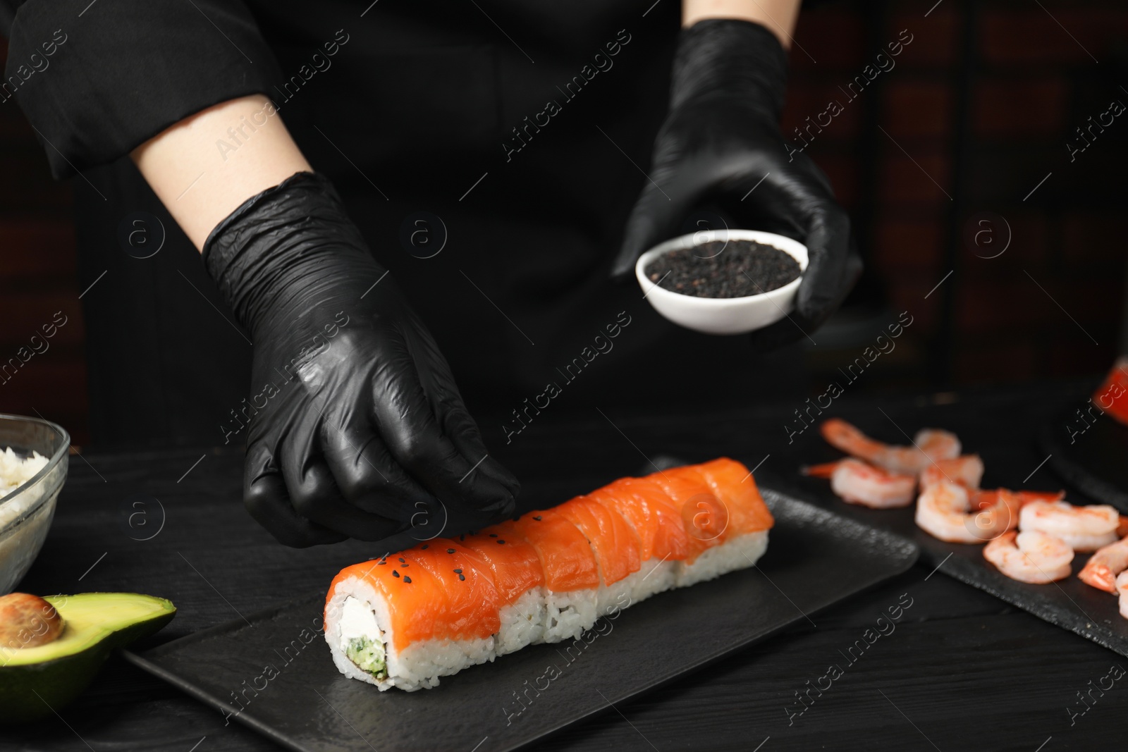 Photo of Chef in gloves adding sesame seeds onto tasty sushi rolls at black wooden table, closeup