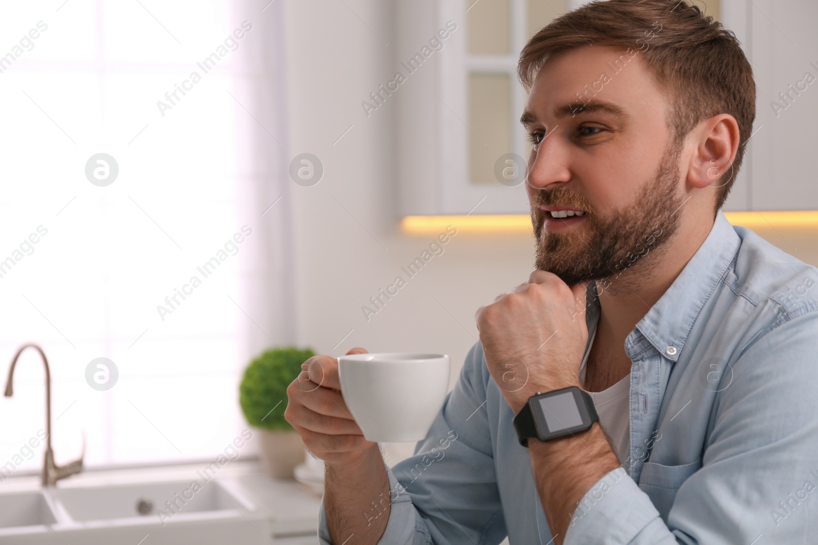 Photo of Young man with smart watch and cup of coffee in kitchen, space for text