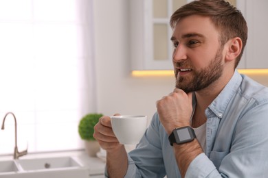 Photo of Young man with smart watch and cup of coffee in kitchen, space for text