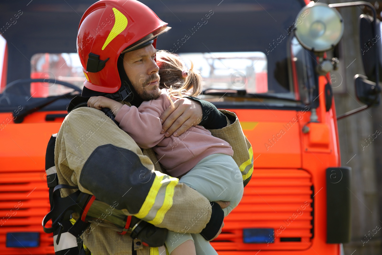 Photo of Firefighter in uniform holding rescued little girl near fire truck outdoors. Save life