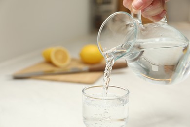 Woman pouring water from jug into glass at white table in kitchen, closeup. Space for text