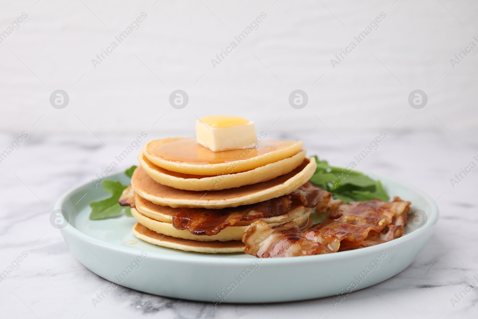 Photo of Tasty pancakes with butter, fried bacon and fresh arugula on white marble table