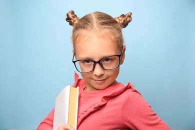 Photo of Cute little girl with glasses and textbook on light blue background