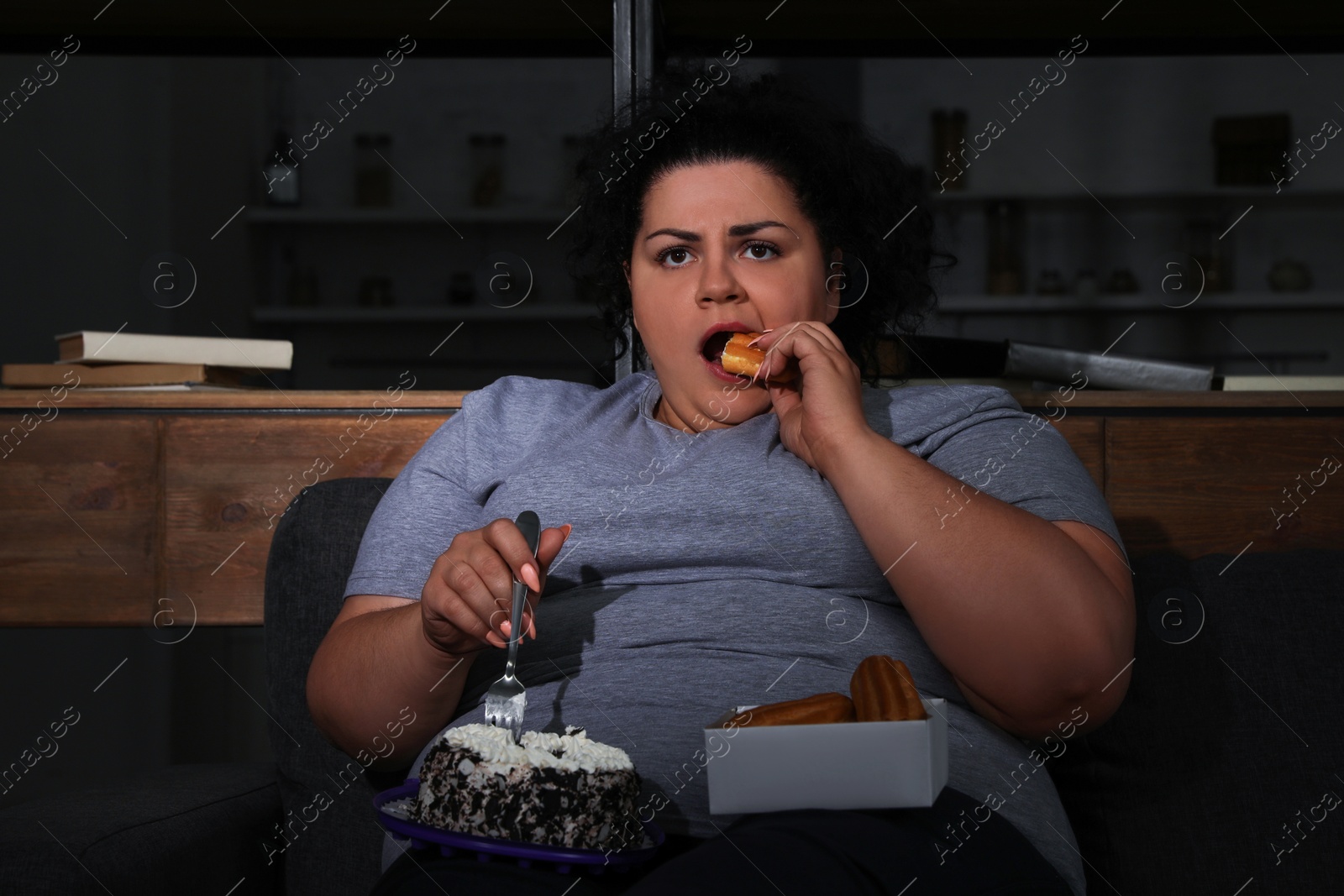 Photo of Depressed overweight woman eating sweets in living room at night