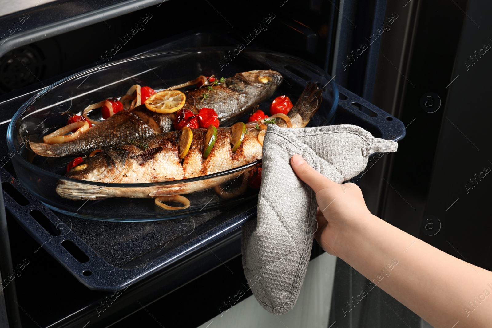 Photo of Woman taking out baking tray with sea bass fish and garnish from oven, closeup