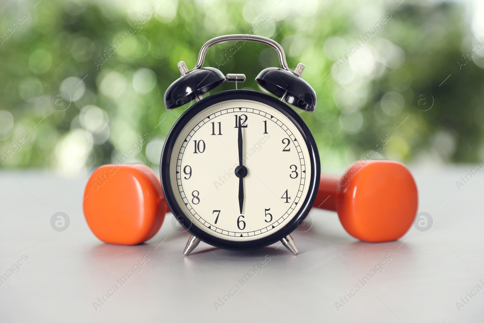 Photo of Alarm clock and dumbbells on grey table against blurred background. Morning exercise