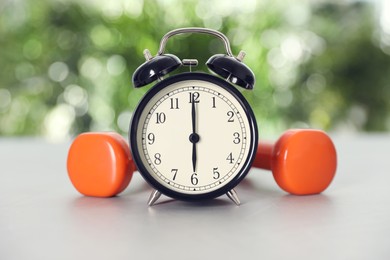 Photo of Alarm clock and dumbbells on grey table against blurred background. Morning exercise