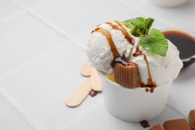 Scoops of tasty ice cream with caramel sauce, mint and candies on white tiled table, closeup. Space for text