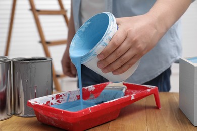 Man pouring light blue paint from bucket into tray at wooden table indoors, closeup