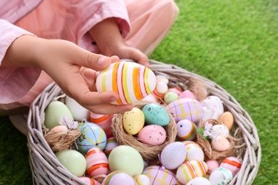 Photo of Child with wicker basket full of Easter eggs on green grass, closeup