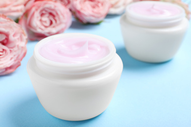 Open jars of cream and flowers on blue background, closeup