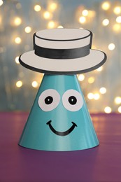 Photo of Funny handmade party hat on purple table against blurred lights