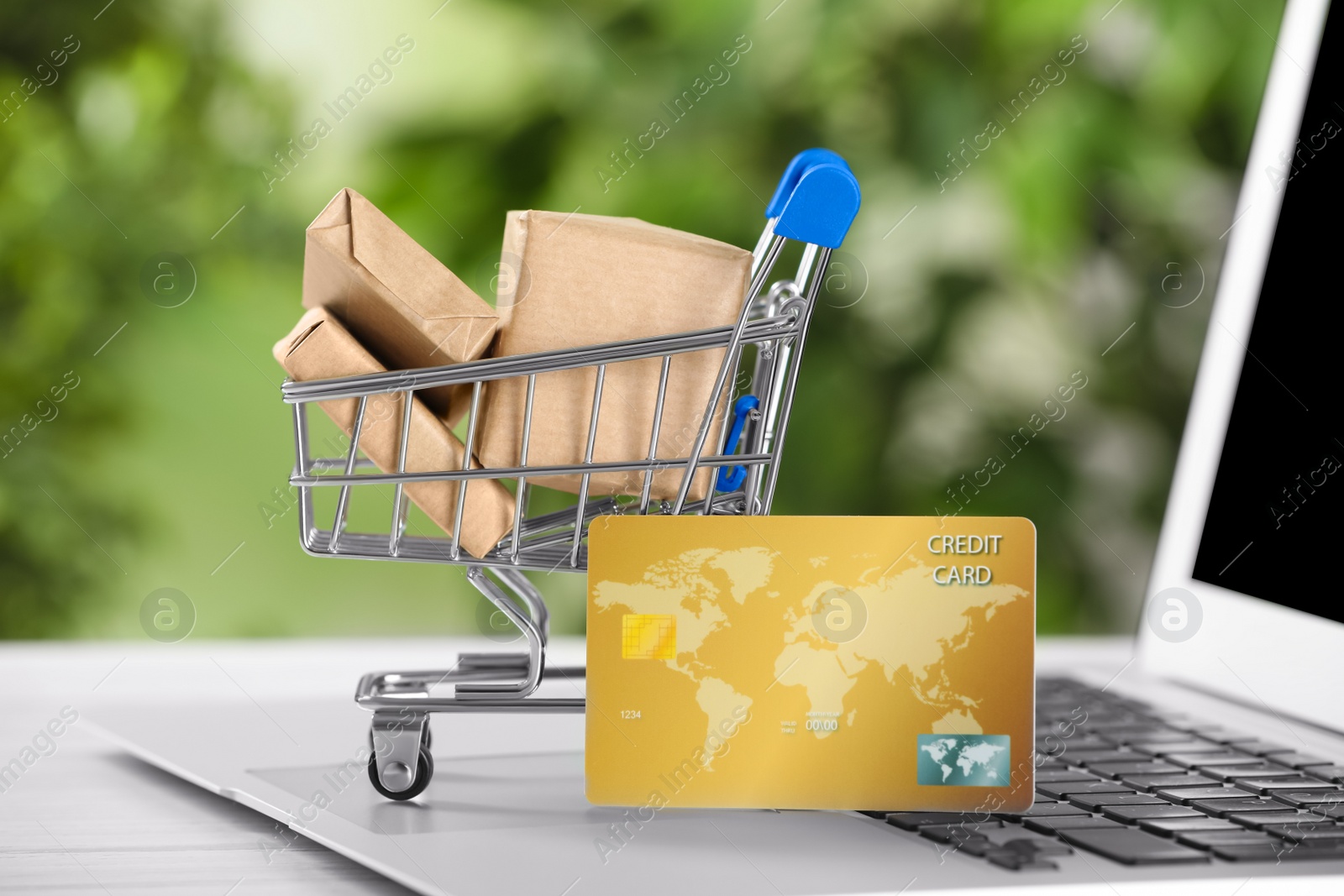 Photo of Online payment concept. Small shopping cart with bank card, boxes and laptop on table, closeup