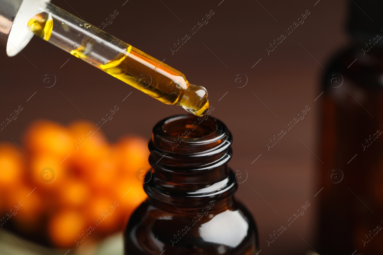 Photo of Natural sea buckthorn oil dripping from pipette into bottle on blurred background, closeup