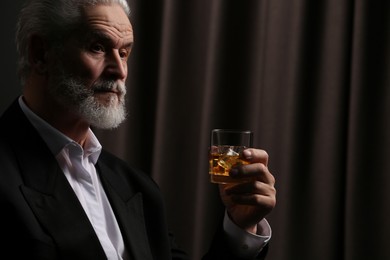 Photo of Senior man in suit holding glass of whiskey with ice cubes on brown background. Space for text