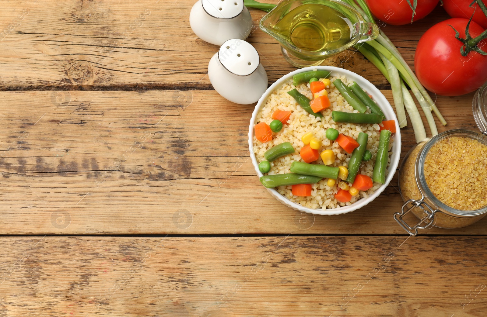 Photo of Delicious bulgur with vegetables in bowl, green onion, oil and tomatoes on wooden table, flat lay. Space for text