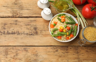 Photo of Delicious bulgur with vegetables in bowl, green onion, oil and tomatoes on wooden table, flat lay. Space for text