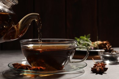 Pouring aromatic anise tea from teapot into glass cup on light grey table