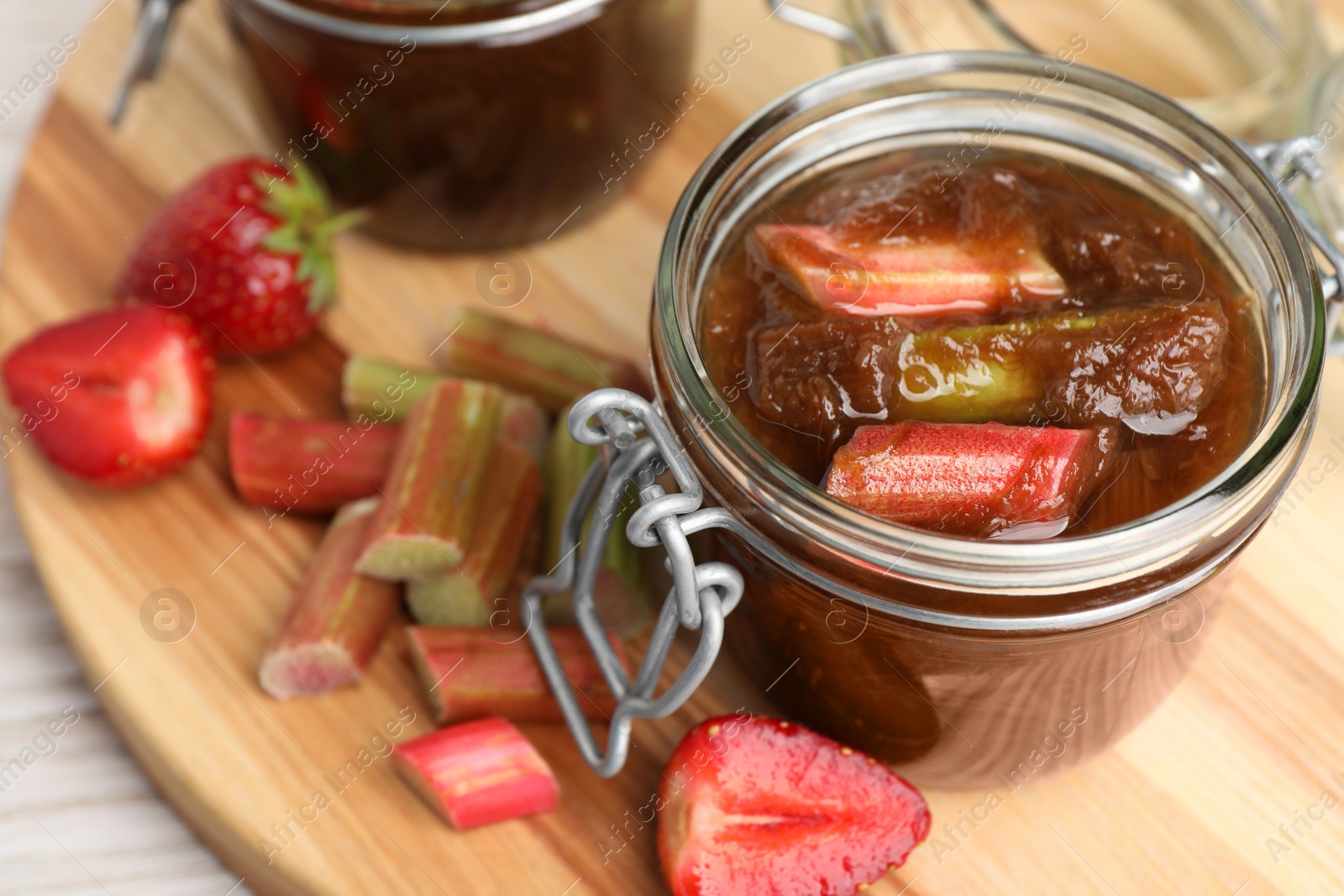 Photo of Jars of tasty rhubarb jam, cut stems and strawberries white wooden table, closeup