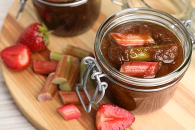 Photo of Jars of tasty rhubarb jam, cut stems and strawberries white wooden table, closeup
