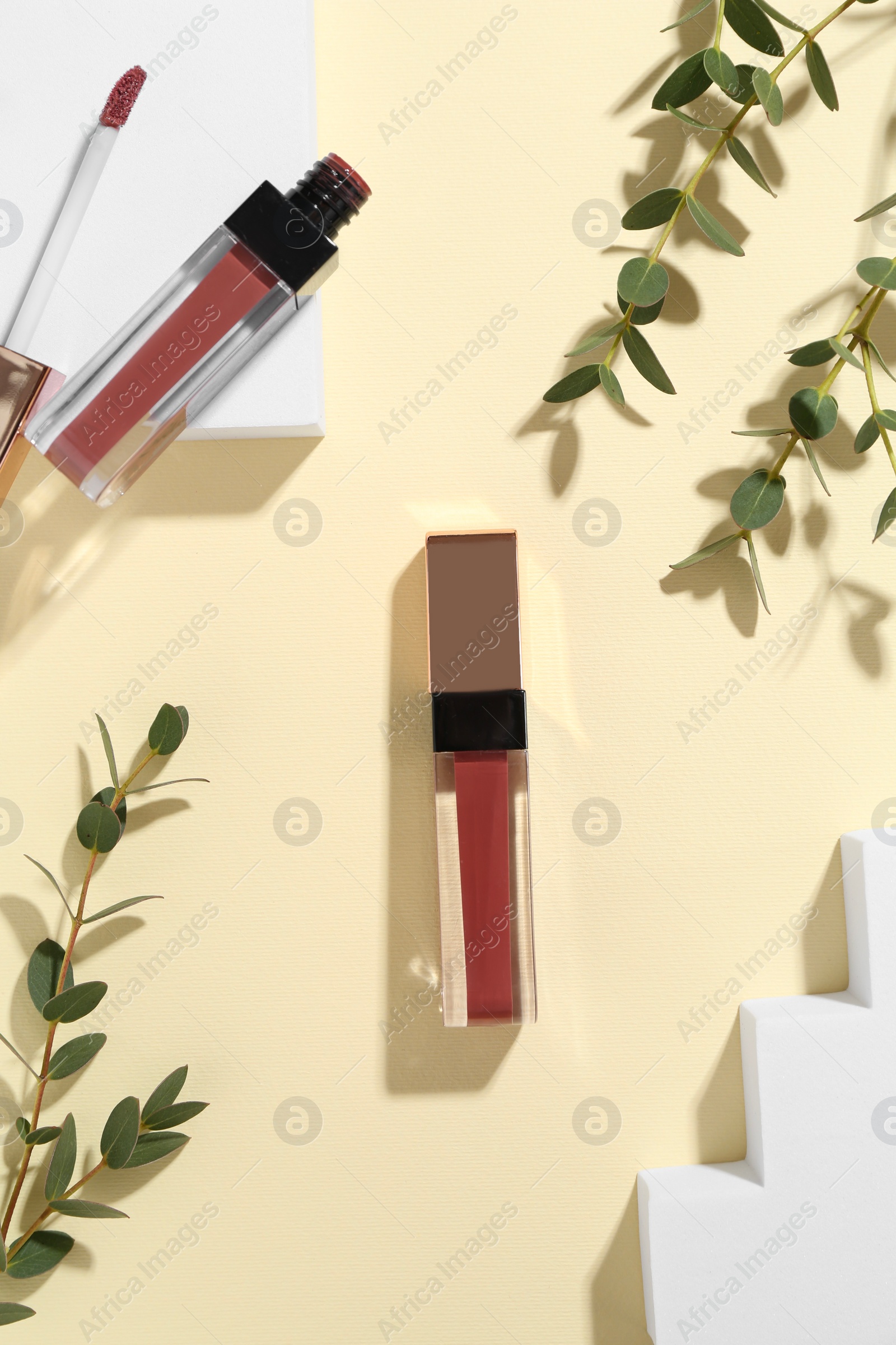 Photo of Lip glosses, podiums and green leaves on pale yellow background, flat lay