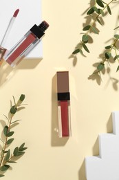 Photo of Lip glosses, podiums and green leaves on pale yellow background, flat lay