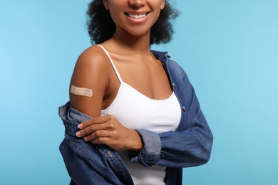 Happy young woman with adhesive bandage on her arm after vaccination against light blue background, closeup