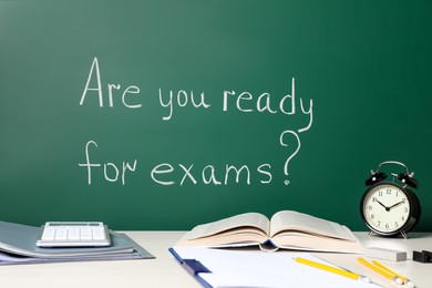 Photo of Different stationery and alarm clock on white table near chalkboard with phrase Are You Ready For Exams